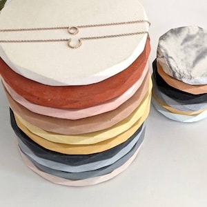 May include: A stack of colourful round concrete coasters with a geometric design. The top coaster is white and has a delicate gold necklace with a small circle pendant on it. A second stack of coasters is made of white, grey, and black marble.