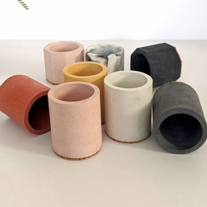 May include: A set of eight small, cylindrical concrete planters in various colours, including black, white, grey, pink, yellow, and orange. Each planter has a cork base.