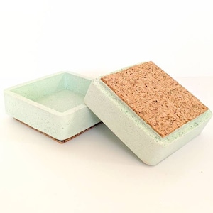 May include: A light green square box with a cork lid. The box is made of concrete and has a speckled texture. The lid has a cork top and a smooth, light green bottom.
