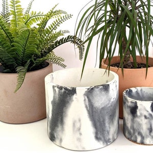Puede incluir: Tres macetas de hormigón de diferentes tamaños con un diseño de mármol. La maceta más grande es blanca con vetas negras, la maceta mediana es marrón y la maceta más pequeña es blanca con vetas negras. Las macetas están vacías.