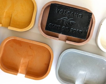 May include: Four concrete soap dishes in different colors: yellow, orange, brown, and gray. A black bar of soap with the text "VOLCANO OLIVE OIL SOAP" is in the brown dish.