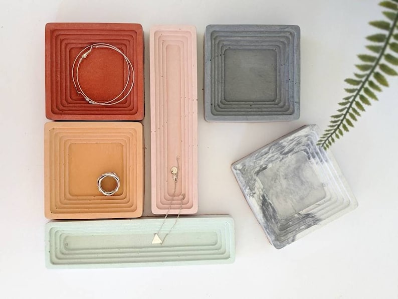 May include: Set of five square concrete trinket dishes with different coloured and textured finishes. The dishes are arranged in a staggered pattern on a white surface.
