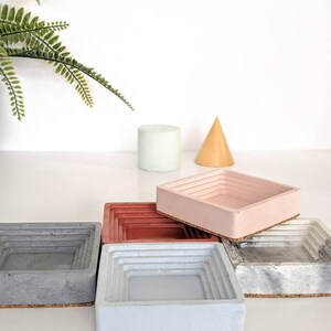 May include: Four square concrete trays with a layered design. The trays are in shades of grey, blue, red, and pink. Each tray has a cork base.