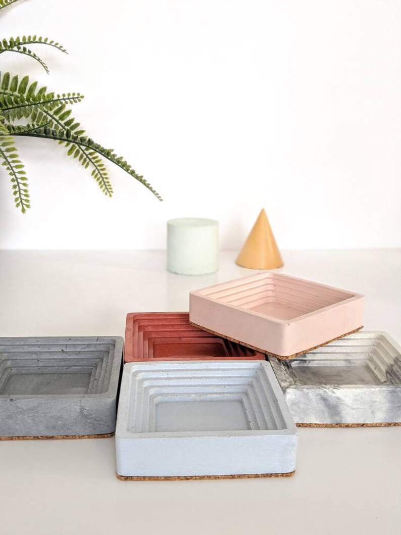 May include: Four square concrete trays with a layered design. The trays are in shades of grey, blue, red, and pink. Each tray has a cork base.