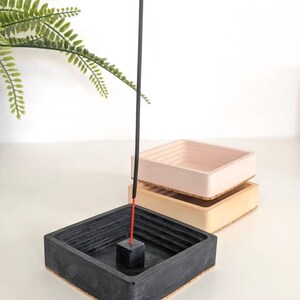 May include: Black, beige, and pink concrete incense holder with a black incense stick. The holder has a square shape with a recessed centre for the incense stick.