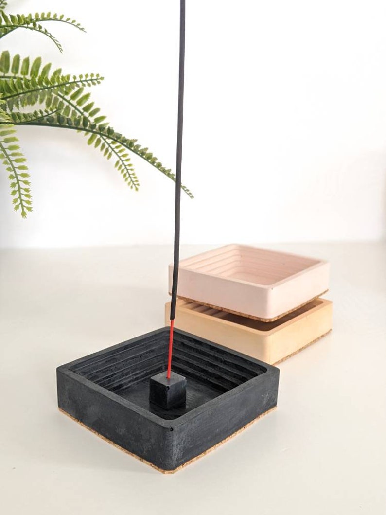 May include: Black, beige, and pink concrete incense holder with a black incense stick. The holder has a square shape with a recessed centre for the incense stick.