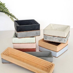 May include: A set of six concrete and wood nesting trays in various colours, including black, white, light blue, light green, pink, and brown. The trays are stacked on top of each other and have a cork base.