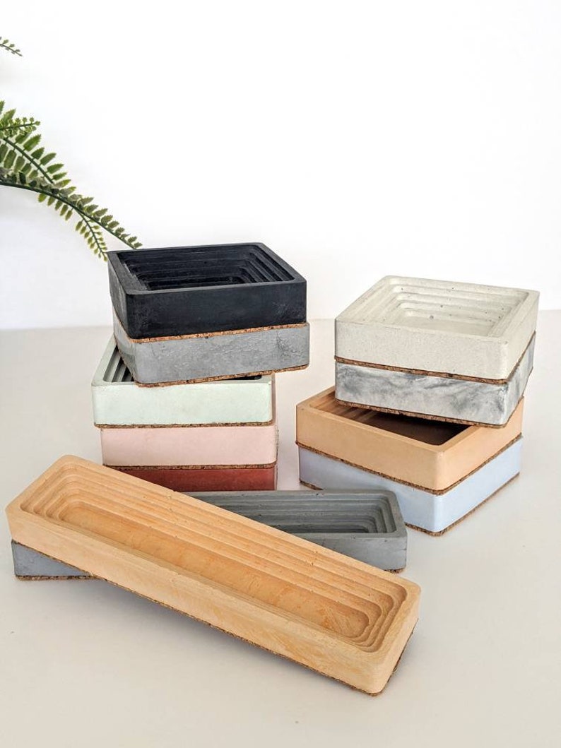 May include: A set of six concrete and wood nesting trays in various colours, including black, white, light blue, light green, pink, and brown. The trays are stacked on top of each other and have a cork base.