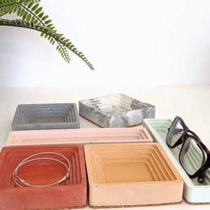 May include: Five concrete trays in different colours, including grey, white, pink, red, and light blue. The trays are stacked on top of each other and have a layered design. A pair of glasses is resting on the light blue tray.