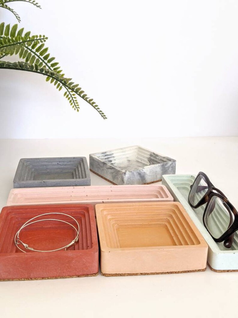 May include: Five concrete trays in different colours, including grey, white, pink, red, and light blue. The trays are stacked on top of each other and have a layered design. A pair of glasses is resting on the light blue tray.