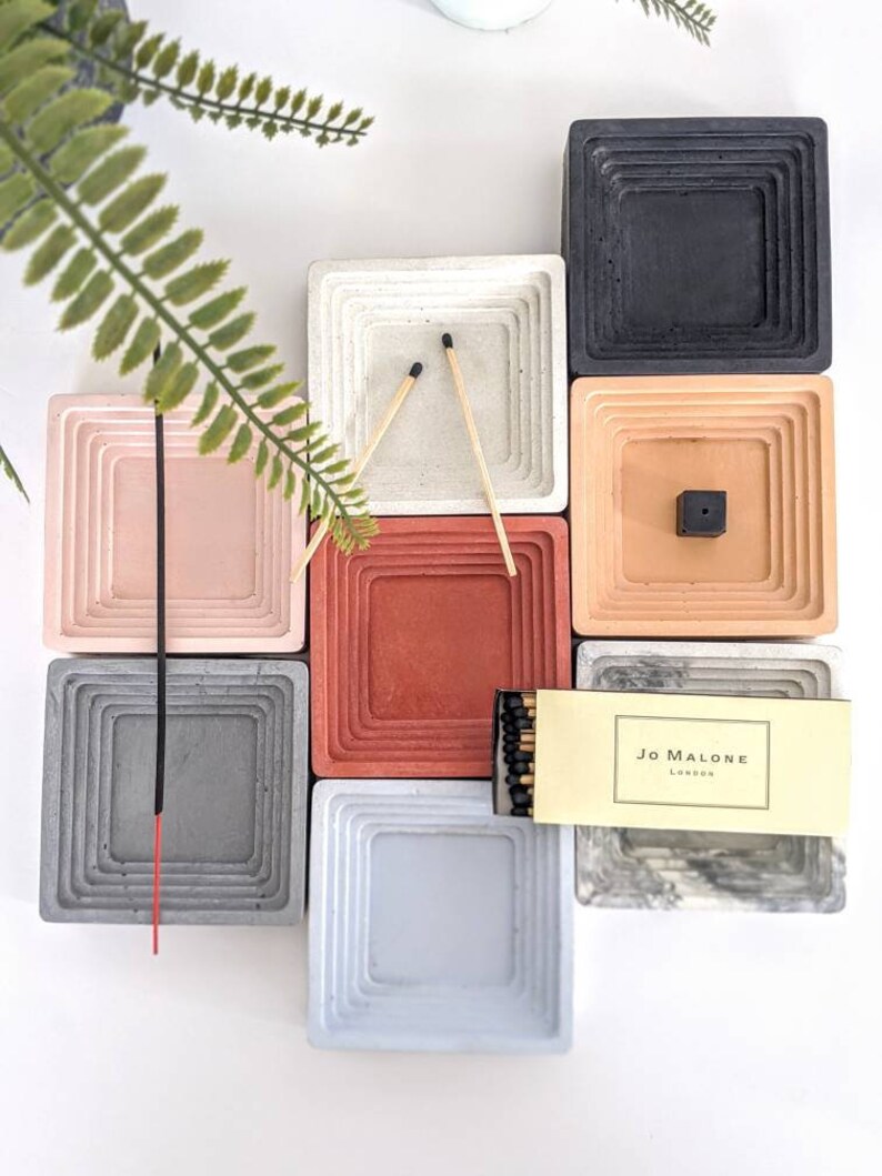 May include: A set of nine concrete incense holders in various colours, including grey, white, pink, red, and beige. The holders are arranged in a grid pattern and feature a layered design. A box of matches with the text "JO MALONE LONDON" is included.