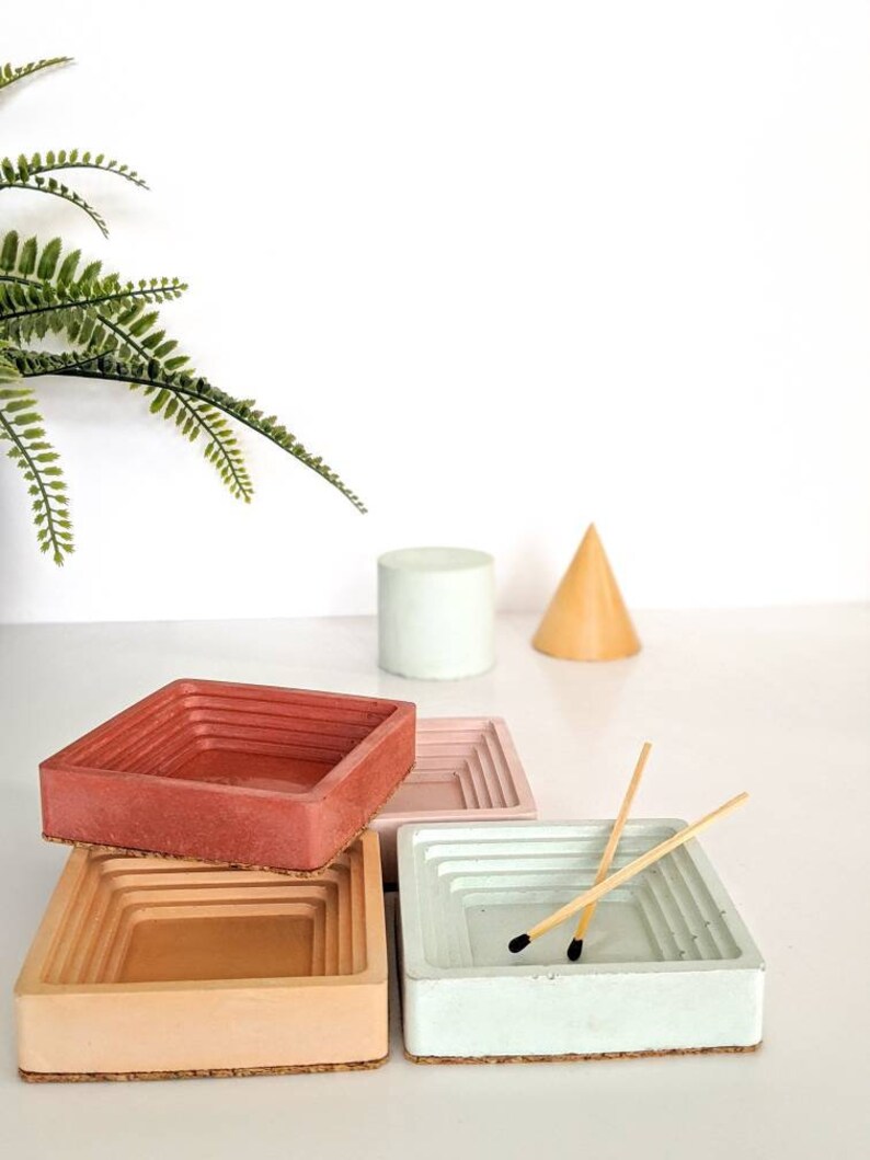 May include: Four square concrete trays in shades of pink, peach, and green. The trays are stacked on top of each other, with the top tray containing two wooden matches.