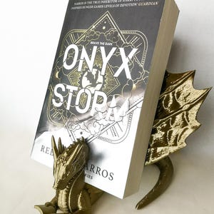 Op de afbeelding: Een gouden drakenboekenlegger die een boek vasthoudt met de titel "Onyx & Storm: Brave the Dark" van Tarros. De boekomslag heeft een zwart en gouden ontwerp met een ster en wolken.