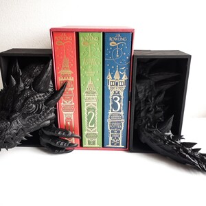 Può includere: Due ferma libri a forma di drago nero con un set di tre libri di Harry Potter. I libri sono in una copertina rossa, verde e blu con lettere dorate. I ferma libri sono in materiale plastico nero e hanno un design di drago dettagliato.