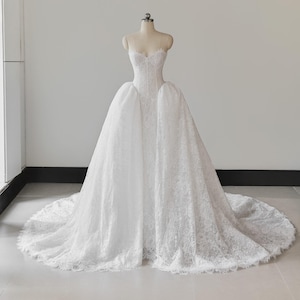 Könnte beinhalten: Ein weißes trägerloses Brautkleid mit einem figurbetonten Oberteil mit herzförmigem Ausschnitt und einem bodenlangen Rock, der mit komplizierter Blumen-Spitze überzogen ist. Das Kleid hat eine strukturierte Silhouette und eine lange Schleppe.