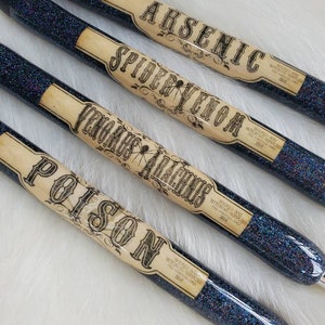 May include: Four black glitter pens with vintage-style labels that read "Arsenic", "Spider Venom", "Venomous Arachnis", and "Poison". Each label has the text "Wicked Way Witching Supplies" and the year "1814".