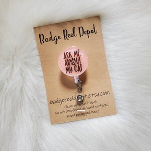 May include: Pink glitter badge reel with a white retractable cord and a round charm that says "Ask Me About My Cat" with paw prints. The charm is attached to the reel with a silver clip. The reel is on a brown card with the text "Badge Reel Depot" and "badgereeldt.etsy.com". The card also includes care instructions: "Clean with soft cloth, Do not drop/bang on hard surfaces, Avoid prolonged heat."