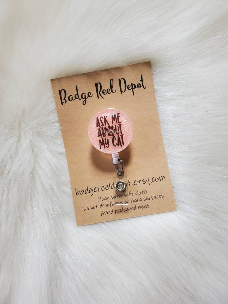 May include: Pink glitter badge reel with a white retractable cord and a round charm that says "Ask Me About My Cat" with paw prints. The charm is attached to the reel with a silver clip. The reel is on a brown card with the text "Badge Reel Depot" and "badgereeldt.etsy.com". The card also includes care instructions: "Clean with soft cloth, Do not drop/bang on hard surfaces, Avoid prolonged heat."