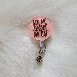 May include: Pink glitter badge holder with a white retractable cord and a black paw print with the text "Ask me about my cat".