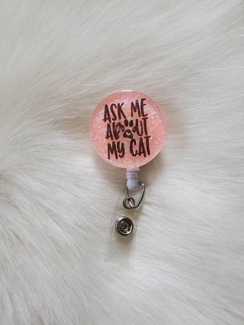 May include: Pink glitter badge holder with a white retractable cord and a black paw print with the text "Ask me about my cat".