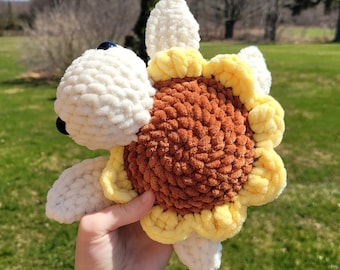 May include: A crocheted sunflower plush toy with a brown center and yellow petals. The sunflower has a white crocheted face with black eyes.
