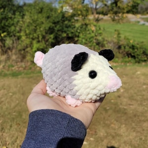 May include: A crocheted opossum toy in grey, cream, and pink. The toy has black ears and eyes. It is held in a hand against a blurred outdoor backdrop. The plushie is a soft, cuddly toy, perfect for children or as a decorative item.