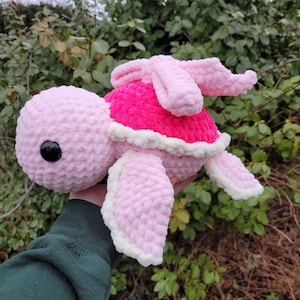 May include: A pink crocheted turtle with a pink and white shell. The turtle has a pink bow on its head and black eyes.
