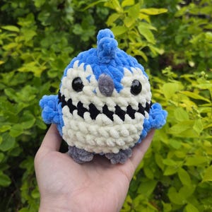 May include: A handmade, plush bluebird toy with a white belly and black accents. The bird has large black eyes and small grey feet. The toy is held in a hand against a backdrop of green foliage.