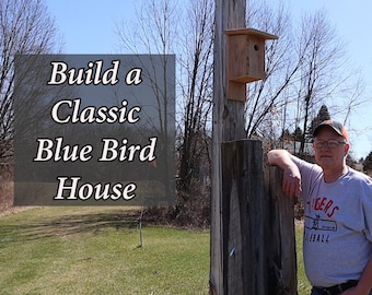 May include: A man stands next to a wooden bluebird house mounted on a wooden post. The house is painted brown and has a small opening for birds to enter. The man is wearing a grey shirt with the word "Tigers" on it. The background is a grassy field with trees in the distance.