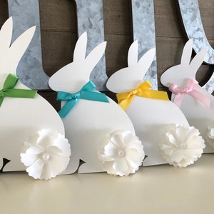 May include: Four white paper bunny cutouts with different colored ribbon bows. Each bunny has a white fabric flower attached to its tail.