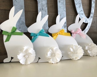 May include: Four white paper bunny cutouts with different colored ribbon bows. Each bunny has a white fabric flower attached to its tail.