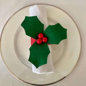 May include: A red and green holly napkin ring with three red berries. The napkin ring is on a white napkin and a white plate with a gold rim.