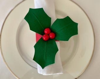 May include: A red and green holly napkin ring with three red berries. The napkin ring is on a white napkin and a white plate with a gold rim.
