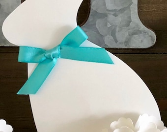 May include: White paper cutout of a bunny rabbit with a turquoise ribbon tied around its neck. The bunny is sitting on a wooden surface with white paper flowers.