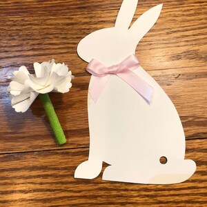 May include: White paper cutout of a bunny with a pink ribbon around its neck. The bunny is sitting on a wooden surface. A white paper flower with a green stem is to the left of the bunny.
