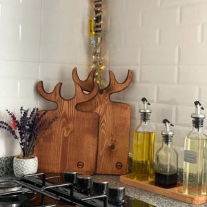 May include: Two wooden chopping boards shaped like deer heads with antlers, featuring a natural brown wood grain finish. Beside them are three glass oil and vinegar dispensers on a wooden tray and a small pot of dried lavender flowers.