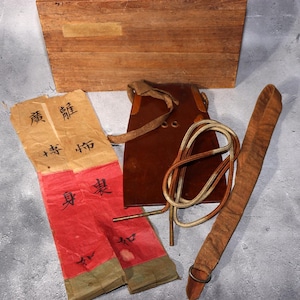 Puede incluir: Un surtido de artículos vintage, que incluyen una caja de madera, una bolsa de cuero con una cuerda y una correa larga de madera con un anillo de metal. También hay dos elementos de papel rojo y beige con caracteres chinos. Los artículos están dispuestos sobre una superficie gris.