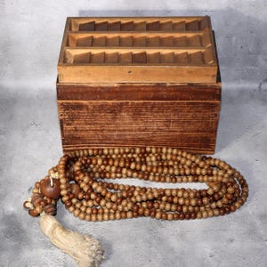 Op de afbeelding: Een houten kist met meerdere compartimenten staat boven een streng houten kralen. De kist is gemaakt van lichtbruin hout met een donkerdere bruine basis. De kralen zijn in verschillende bruin- en beigetinten, met een kwast.