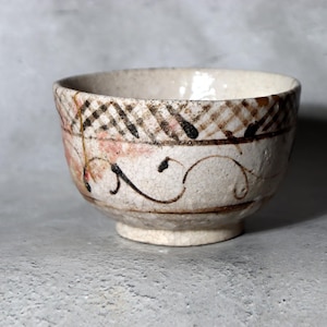 May include: A small, off-white ceramic bowl with a textured surface. The bowl features a decorative pattern of brown and black crosshatching, pink accents, and swirling black lines. The bowl sits on a small, circular base.