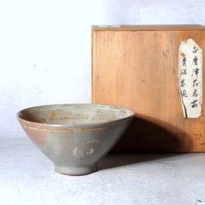 May include: A pale gray ceramic bowl with a circular design on the side. The bowl has a wide opening and a slightly flared base. A wooden box is in the background with Japanese text on a label.