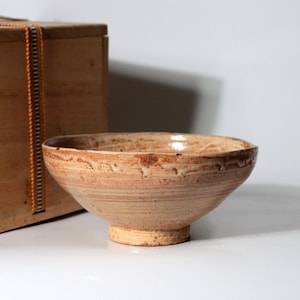 May include: A shallow, wide-rimmed ceramic bowl with a textured, light tan and earthy brown glaze. The bowl features a tapered base and a slightly uneven, organic rim. It is positioned in front of a rectangular wooden storage box with a vertical orange strap.