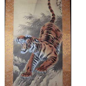 May include: A vertical wall hanging scroll featuring a painting of a snarling tiger with orange, black, and white fur. The tiger is positioned in a pouncing stance against a backdrop of bamboo. The scroll has decorative gold patterned borders.