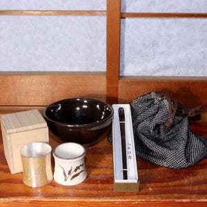 May include: A set of Japanese tea ceremony items including a brown tea bowl, two small ceramic cups, a wooden box, a black and white patterned fabric bag, and a wooden stick with a black handle.
