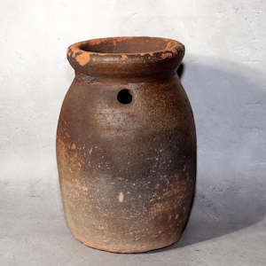 以下が含まれることがあります： 丸みを帯びたボディとわずかに広がった縁を持つ、素朴な茶色の陶器の壺。上部に小さな丸い穴があり、使い古された外観です。色は茶色と赤みがかったオレンジ色の混合です。