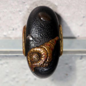 Peut inclure: Objet noir de forme ovale avec une surface texturée. Il présente des éléments décoratifs dorés et rouges, dont un coquillage stylisé et des bandes dorées. Un petit accent brun foncé est visible en haut.