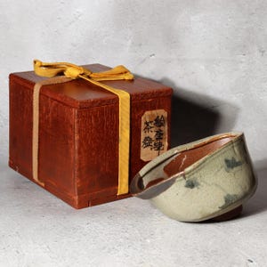 Pode incluir: Uma caixa de madeira castanha escura, amarrada com uma fita amarela, ao lado de uma tigela de chá de cerâmica. A tigela de chá é verde clara com detalhes em castanho e azul. A caixa tem um rótulo.