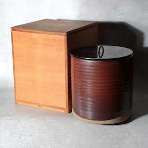 May include: A cylindrical ceramic jar with a dark reddish-brown glazed finish and a smooth black lid featuring a small central handle, positioned next to a square wooden storage box with a flat lid and visible wood grain texture.