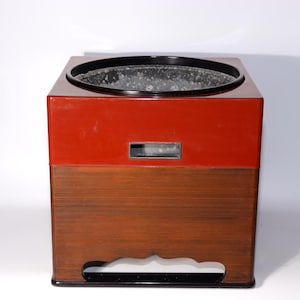 May include: A square, lacquered wooden container with a red upper section and a lower section with horizontal wood grain. The top has a black rim and a circular opening. A rectangular opening is visible on the front.
