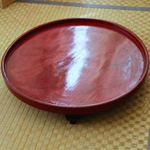 May include: A round, red lacquered tray with a slightly raised edge. The tray has a glossy finish and appears to be made of wood. The tray is sitting on a woven mat.