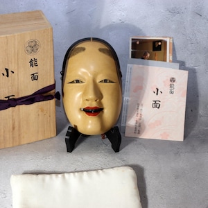 以下が含まれることがあります： 穏やかな表情の伝統的な日本の能面。黒髪、繊細なペイントされた顔の特徴、赤い唇が特徴です。マスクは黒いスタンドに展示され、木箱と情報資料が付属しています。